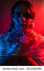 Handsome Young Man With Long Hair Wrapped In A Plastic Bag. Man Covered With Cellophane Wrap. Portrait In Blue And Red Lighting.