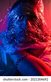 Handsome Young Man With Long Hair Wrapped In A Plastic Bag. Man Covered With Cellophane Wrap. Portrait In Blue And Red Lighting.