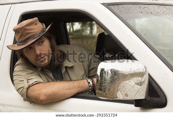 Handsome young man with long\
hair in brown cowboy hat driving white 4x4 car after rain. Safari\
style. 