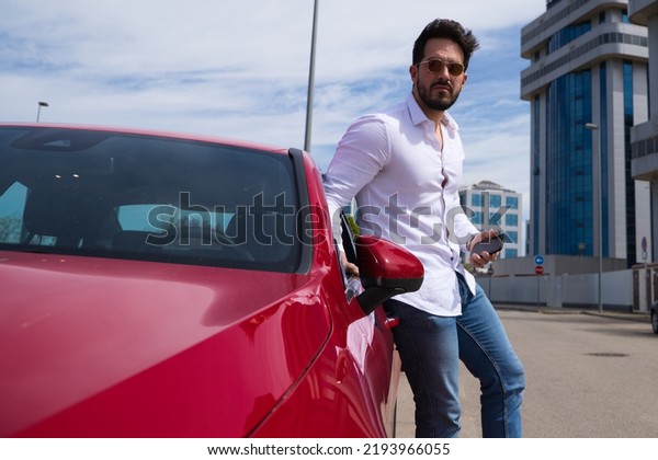 Handsome young man, looking at his mobile\
phone next to his red sports car. The man is wealthy. High standard\
of living and well positioned\
financially.