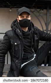 Handsome young man model with black cap and protective medical mask in black jacket and hoodie sits on the street