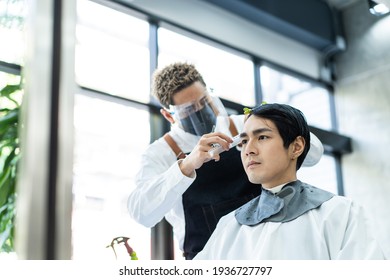 Handsome Young Man In Modern Salon Getting A Haircut Service By A Stylist Asian Male Hairdresser Wearing A Face Shield And Mask For Protection And Practicing Social Distance During Covid 19 Pandemic.