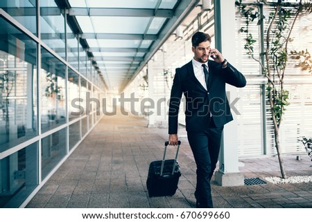 Handsome young man on business trip walking with his luggage and talking on cellphone at airport. Travelling businessman making phone call.