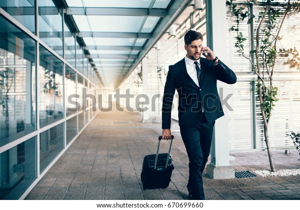 Handsome young man on business trip walking with\
his luggage and talking on cellphone at airport. Travelling\
businessman making phone\
call.