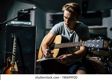 Handsome Young Man On Rehearsal Base. Lyric Singer With Acoustic Guitar Is Writing Notes And Lyrics In Notebook.