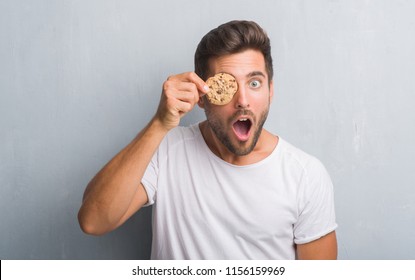 Handsome young man over grey grunge wall eating chocolate chip cooky scared in shock with a surprise face, afraid and excited with fear expression