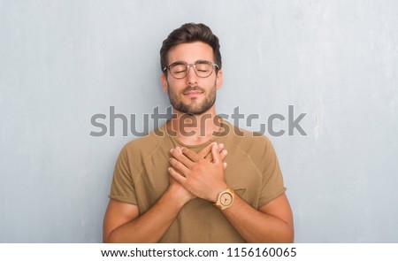 Handsome young man over grey grunge wall wearing glasses smiling with hands on chest with closed eyes and grateful gesture on face. Health concept.