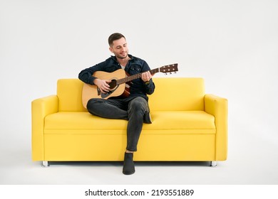 Handsome young man playing acoustic guitar whlile sitting on yellow sofa over white background. Hobby, music concept - Powered by Shutterstock