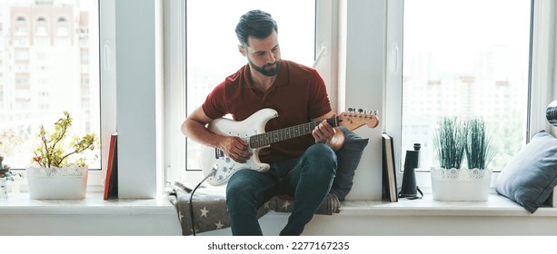 Handsome young man playing acoustic guitar while relaxing on the window sill at home - Powered by Shutterstock