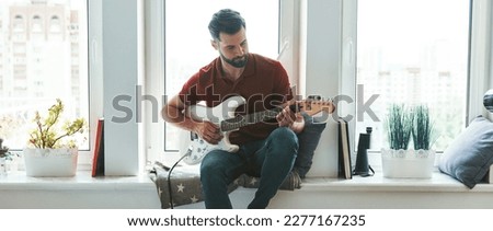 Handsome young man playing acoustic guitar while relaxing on the window sill at home