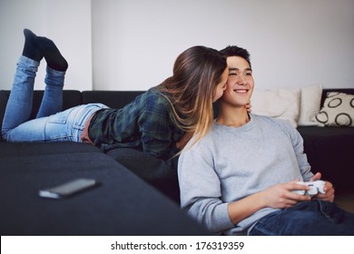 Handsome Young Man Playing Video Game While His Girlfriend Whispering Something In His Ears. Teenage Asian Couple Relaxing In Living Room At Home.