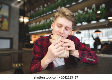 A handsome young man with pleasure bites an appetizing sandwich in a cafe. A student dishes a sandwich in a cafe