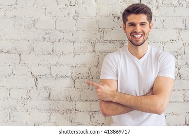 Handsome young man is pointing away, looking at camera and smiling, standing against white brick wall - Powered by Shutterstock