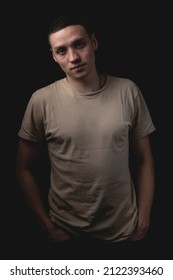 Handsome Young Man Posing Over Black Background. Studio Photo With One Light Source. A Serious Guy Stands With His Hands In His Pockets.