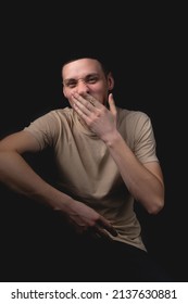 Handsome Young Man Posing Over Black Background. Studio Photo With One Light Source. Smiling Guy Cheerfully Posing In The Studio. The Guy Covers His Face With His Hand, Yawning.