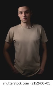 Handsome Young Man Posing Over Black Background. Studio Photo With One Light Source. A Serious Guy Stands With His Hands In His Pockets.