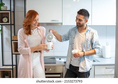 Handsome young man pouring hot water from electric kettle into his redhead girlfriend's cup in the kitchen.