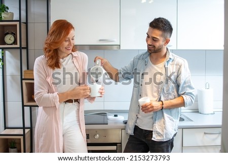 Handsome young man pouring hot water from electric kettle into his redhead girlfriend's cup in the kitchen.