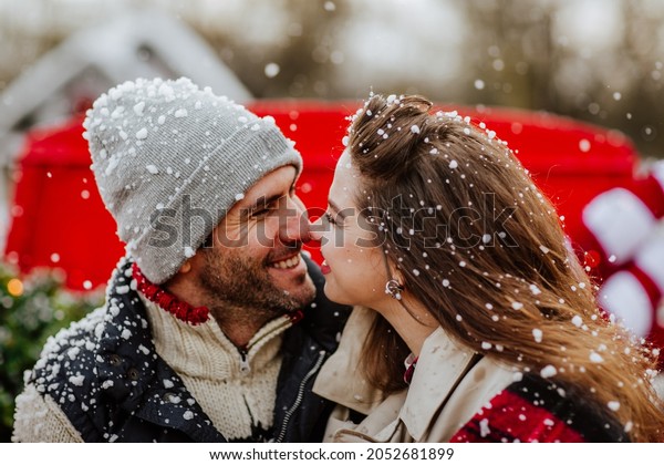 Handsome young\
man and pretty curly haired woman in winter clothes posing in red\
Christmas car with decor against white house and fence. Focus is at\
the woman. Snowing. Holiday\
concept.