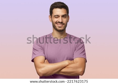 Handsome young man in purple t-shirt, with crossed arms smiling and winking, looking at camera isolated on purple background