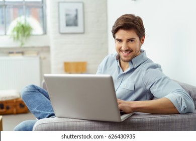 Handsome Young Man Relaxing At Home With A Laptop Computer Balanced On The Arm Of The Sofa Looking At The Camera With A Warm Friendly Smile