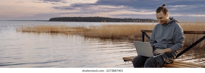 A handsome young man is resting and working on a laptop near the sea. A freelancer works sitting on a wooden bench on a pier by the water. Banner for website header design with copy space. - Powered by Shutterstock