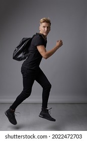 Handsome Young Man Running With A Backpack On Shoulders Smiling, Moving Hands And Looking Back On Studio Light Gray Background. Stylish Man With Fashionable Hairdo.