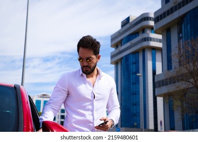 Handsome Young Man, Sculpted Body Next To His Red Sports Car. The Man Is Wealthy And Dressed In Modern Clothes. Sports Car