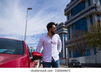 Handsome Young Man, Sculpted Body Next To His Red Sports Car. The Man Is Wealthy And Dressed In Modern Clothes. Sports Car