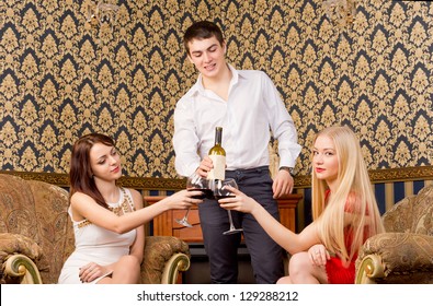 Handsome young man serving wine to two glamorous young ladies seated together in vintage armchairs