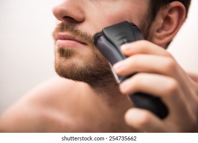 Handsome young man is shaving with electric razor while looking at the mirror.