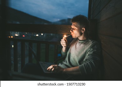 Handsome young man sits at night on the balcony with a laptop on his lap and lights a cigarette with a lighter.Evening portrait freelancer smokes and works on a laptop on a balcony in an apartment