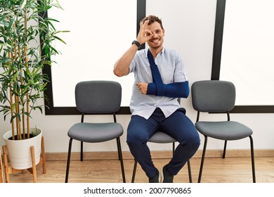 Handsome Young Man Sitting At Doctor Waiting Room With Arm Injury Doing Ok Gesture With Hand Smiling, Eye Looking Through Fingers With Happy Face. 