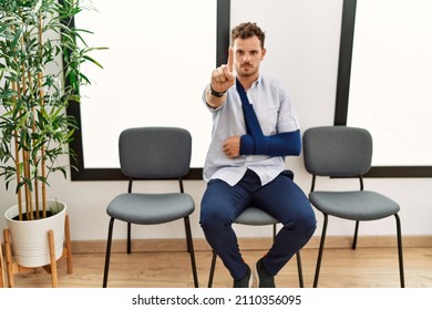 Handsome Young Man Sitting At Doctor Waiting Room With Arm Injury Pointing With Finger Up And Angry Expression, Showing No Gesture 