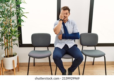 Handsome Young Man Sitting At Doctor Waiting Room With Arm Injury Covering One Eye With Hand, Confident Smile On Face And Surprise Emotion. 