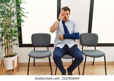 Handsome Young Man Sitting At Doctor Waiting Room With Arm Injury Doing Ok Gesture Shocked With Surprised Face, Eye Looking Through Fingers. Unbelieving Expression. 