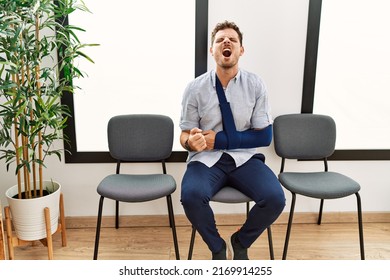 Handsome Young Man Sitting At Doctor Waiting Room With Arm Injury Angry And Mad Screaming Frustrated And Furious, Shouting With Anger. Rage And Aggressive Concept. 