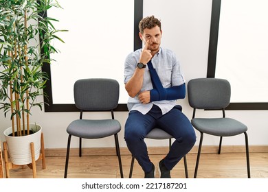 Handsome Young Man Sitting At Doctor Waiting Room With Arm Injury Pointing To The Eye Watching You Gesture, Suspicious Expression 