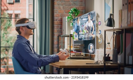 Handsome Young Man Sitting In Loft Living Room At Home, Using Virtual Reality Headset With Controllers To Check Social Media Streaming Application. Male Is Watching A Cooking TV Show.