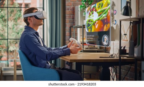 Handsome Young Man Sitting In Loft Living Room At Home, Using Virtual Reality Headset With Controllers To Check Social Media Streaming Application. Male Is Watching A Cooking TV Show.