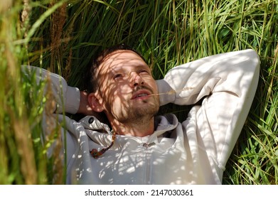 A handsome young man with a smile looks away lying in the tall grass, which casts a shadow from the bright sun on his face