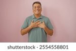 Handsome young man smiling with eyes closed and hands on heart, standing over an isolated pink background in a soft green shirt, embodying gratitude and joy in a candid portrait