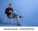 Handsome young man smiling and using social media over smart phone on chair against blue background