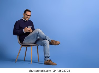 Handsome young man smiling and using social media over smart phone on chair against blue background - Powered by Shutterstock