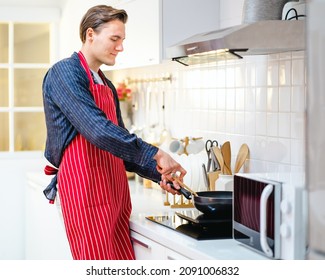 Handsome Young Man Is Standing At Cabinet In Kitchen Looks At Frying Pan On The Stove. Healthy Male Chef Wears Red Apron Preparing Food For Dinner Or Lunch. Cheerful Guy Enjoys Cooking Alone At Home. 