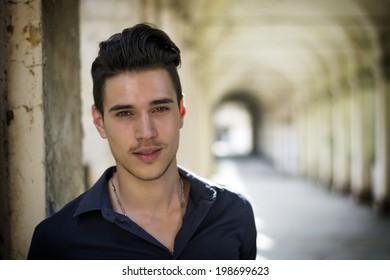 Handsome young man standing outdoors under old colonnade in european town, smiling