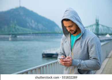 A handsome young man standing in training clothes and using his phone with earphones at the riverside
 - Powered by Shutterstock