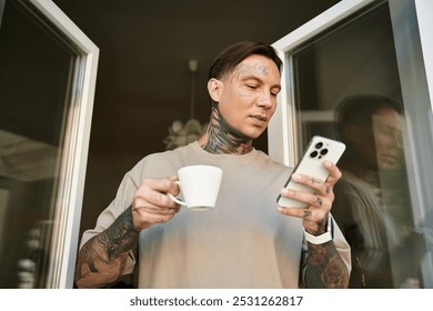 A handsome young man stands on his balcony, sipping coffee and browsing his phone, relaxed at home. - Powered by Shutterstock