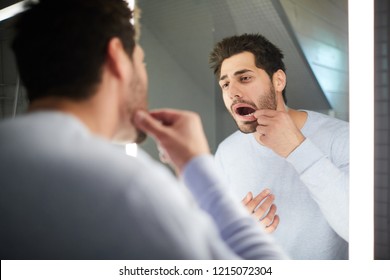 Handsome Young Man With Stubble Keeping Mouth Open While Checking Tooth And Looking Into Mirror In Bathroom