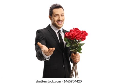 Handsome young man in a suit holding a bunch of red roses and showing at the camera with his hand isolated on white background - Powered by Shutterstock